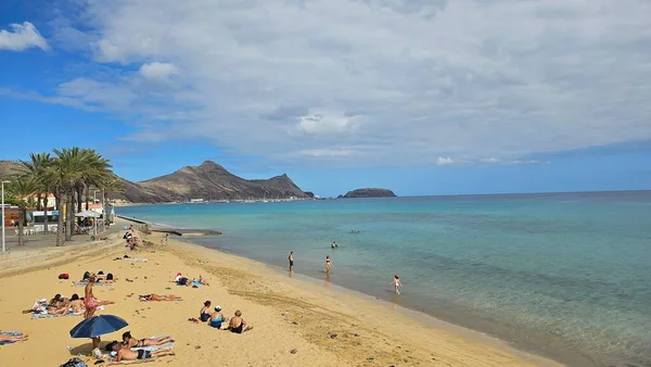 Portekiz 'in Madeira kentindeki Porto Santo plajının panoramik manzarası, altın kum, turkuaz Atlantik suyu, rahat sahil ziyaretçileri, palmiye çizgili gezinti güvertesi ve dokulu bulutların altındaki volkanik ada manzarası..