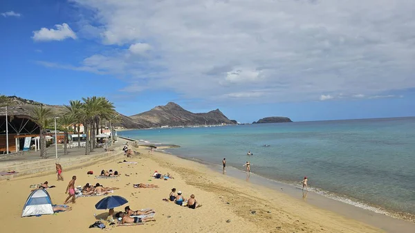 Madeira, Portekiz 'deki güneşli Porto Santo plajı, altın kum, turkuaz Atlantik suyu, dinlenen ve yüzen plajcılar, palmiye çizgili gezinti güvertesi ve dramatik bulutlar altında uzak volkanik ada manzarası..