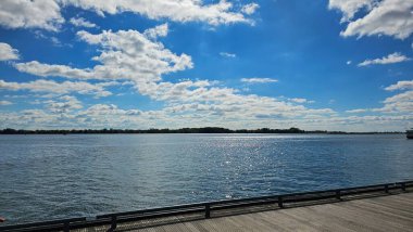 Parlak rıhtım manzarası, sakin su üzerinde ışıl ışıl güneş ışığı, ağaçlarla kaplı uzak kıyı şeridi, ve açık bir günde ahşap bir iskeleden seyredilen canlı mavi gökyüzü boyunca dağınık beyaz bulutlar..