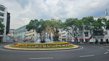 Orta Funchal, Madeira, Portekiz 'de şehir sokakları, dükkanlar ve bulutlu bir gökyüzü altında modern binalarla çevrili çiçek peyzajlı bir çeşme. 21 Eylül 2024