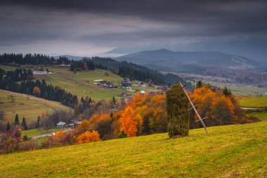 Saman yığını, yuvarlanan tepeler ve dramatik bulutlu gökyüzünün altındaki köy, renkli sonbahar yaprakları ve arka planda dağ manzarası olan sonbahar kırsalları