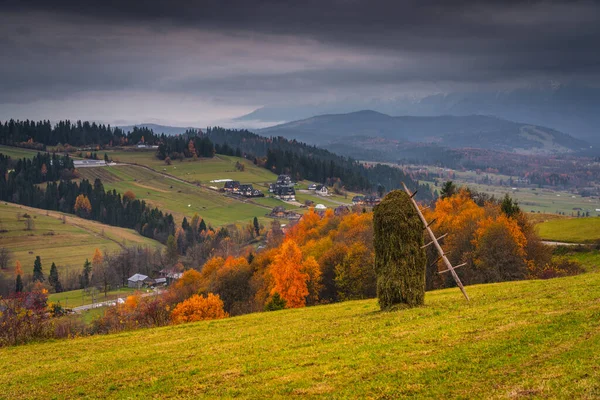 Saman yığını, yuvarlanan tepeler ve dramatik bulutlu gökyüzünün altındaki köy, renkli sonbahar yaprakları ve arka planda dağ manzarası olan sonbahar kırsalları