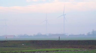Tarımsal alanların üzerinde duran rüzgar türbinleri düşük görüş sisi, ilk saatlerde sessiz renklerle yakalanmış, ölçek, mesafe ve atmosfer derinliğini vurguluyor.