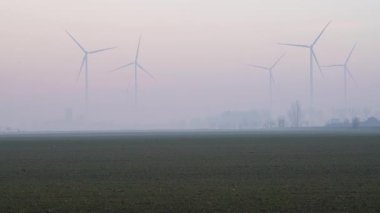 Ufuktaki rüzgar türbinleri sakin bir kırsal ovada, sis ve pastel gökyüzü tonlarıyla yumuşatılmış, sakin ve sürdürülebilir bir enerji manzarası sunar.