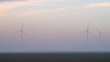 Uzak rüzgar çiftliği şafak vakti yoğun sis yüzünden kısmen kararıyor. Yenilenebilir enerji altyapısının yumuşak ufka karıştığı sakin bir kırsal çevre sunuyor.