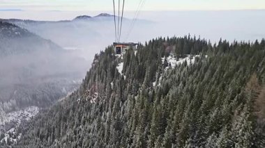 Dağ teleferiği istasyonu, bulutların üzerindeki orman zirvesi, katmanlı tepeler, kar bantları ve derinlik ve sinematik manzara yaratan sis içeren kış alp manzarası.