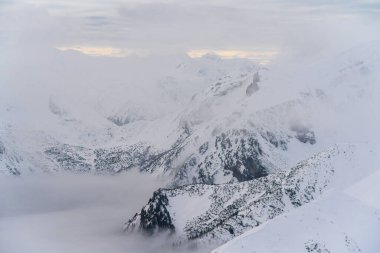 Yoğun bir bulut denizi üzerinde keskin tepelerin yükseldiği, yumuşak ışığın katmanlı zirveleri ve derin vadileri kısmen sürüklenen sis tarafından yuttuğu dramatik bir kış manzarası, Tatras.