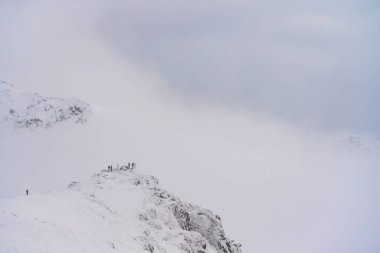 Dar, karla kaplı bir tepe, sisli bir denizin üzerinde yükseliyor ve küçük şekiller, ölçekleri vurguluyor. Yumuşak kış ışığı ve minimum bileşim sakin, alp bir atmosfer yaratır..
