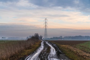 Yüksek voltaj iletim kulesine giden kırsal toprak yol, puslu sabah koşulları, enerji altyapısı, elektrik taşımacılığı ve kırsal ölçekte vurgulanan dramatik gökyüzü