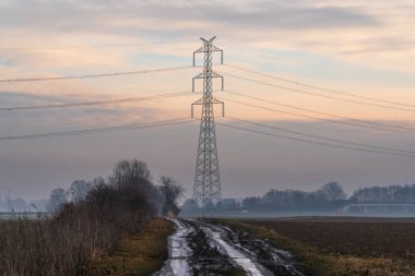 Yüksek voltaj iletim kulesine doğru giden kırsal yol, puslu sabah koşulları, enerji altyapısı, elektrik taşımacılığı ve kırsal ölçekte vurgulanan dramatik gökyüzü
