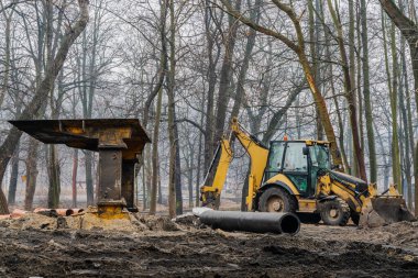 Altyapı inşaatı sırasında çamurlu orman arazisinde çalışan sarı kazıcı, toprak kazan ağır makineler ve kış koşullarında yeraltı boru hattı tesisatı için hazırlık sahası