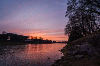 Uzh Nehri, Ukrayna 'nın Uzhhorod kentinden akar. Dağınık bulutlar ve kıyı boyunca uzanan siluetli ağaçlarla dramatik bir günbatımı gökyüzünün sıcak tonlarını yansıtır.