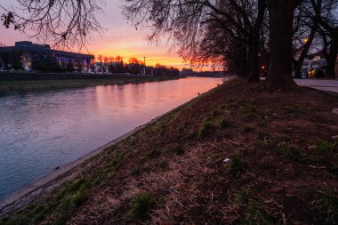 Uzh Nehri Ukrayna 'nın Uzhhorod kentinden akar, siluetli ağaçların ve binaların arkasında dramatik bir günbatımının sıcak tonlarını yansıtır.