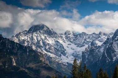 Parçalı bulutlu bir gökyüzünün altında, engebeli, karla kaplanmış zirveleriyle dramatik dağ manzarası Polonya Tatralarının doğal güzelliklerini gözler önüne seriyor.