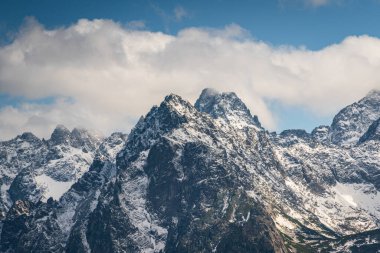Parçalı bulutlu bir gökyüzünün altında, engebeli, karla kaplanmış zirveleriyle dramatik dağ manzarası Polonya Tatralarının saf güzelliğini gözler önüne seriyor.