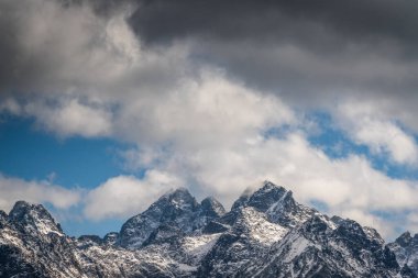 Engebeli, karla kaplı dağ zirveleri dönen bulutlar ve mavi parçalarla dolu dramatik bir gökyüzünü delip geçer.