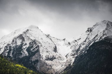 Slovakya 'daki Belianske Tatraları' nın görkemli karlı zirveleri dramatik bulutlu gökyüzünün altındaki koyu yeşil ormanlarla tezat oluşturuyor.