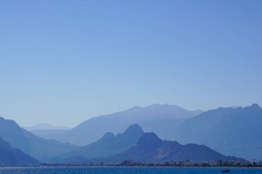 Puslu mavi bir dağ manzarası ufuk boyunca uzanır, sakin denizle ve uzak bir şehrin siluetiyle karşılaşır.