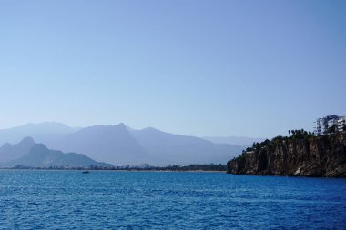 Türkiye 'nin Antalya kenti yakınlarındaki Akdeniz kıyılarının geniş bir görüntüsünde engebeli uçurumlar, uzak bir şehir silueti ve berrak mavi gökyüzünün altındaki puslu dağlar görülüyor.