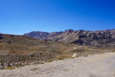 Issız ve çorak bir dağ sırası seyrek bitki örtüsü ve geniş bulutsuz gökyüzünün altındaki araziye uzanan dolambaçlı toprak bir yol.