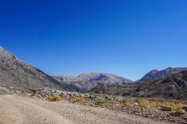 Tozlu toprak bir yol kayalık, kurak bir vadiden uçsuz bucaksız, bulutsuz bir gökyüzünün altındaki uzak, görkemli dağ sıralarına doğru esiyor.