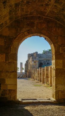 Yıpranmış sütunları ve çökmüş duvarları olan eski bir Akdeniz yapısının güneş ışığıyla aydınlanmış kalıntılarına bakan yıpranmış bir taş kemer yolu.