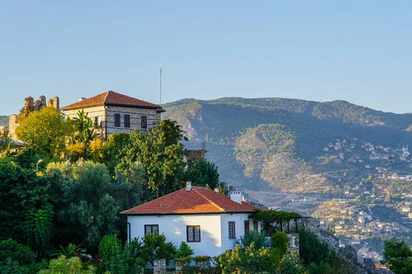 Kırmızı kiremit çatılı geleneksel Türk evleri, uzak bir kasaba ve açık mavi gökyüzü altındaki yemyeşil dağlara bakan yemyeşil bir yamaçta yer alıyor.