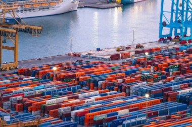 Geniş renkli nakliye konteynırları büyük bir yolcu gemisinin yanındaki hareketli bir liman terminaline düzgünce istiflenmiştir.