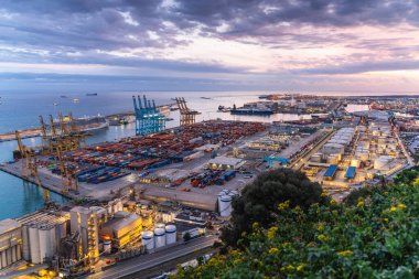 Alacakaranlıktaki hareketli bir limanın havadan görünüşü, engin konteynır yığınlarını, endüstriyel tesisleri ve dramatik bir gökyüzüne karşı büyük vinçleri gösteriyor.