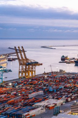 Büyük, aktif bir nakliye limanının havadan görünüşü. Büyük vinçler ve sönen ışığın aydınlattığı sayısız renkli konteynırlar.