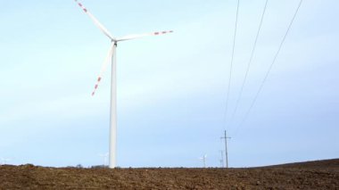 Görkemli rüzgâr türbinleri ekili bir alanda dimdik ayakta duruyor. Arkaplandaki elektrik hatlarına ve dingin gökyüzüne karşı yenilenebilir enerji sağlıyor.