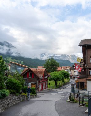 Geleneksel ahşap evleri ve arka planda modern bir binası olan bir adam İsviçre köyünde bir caddede yürüyor. Dağlar bulutlarla örtülmüştür..