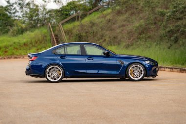 Mavi BMW M3 yan profil görünümü, asfalt zeminde gösterişli performans arabası doğa arka planı