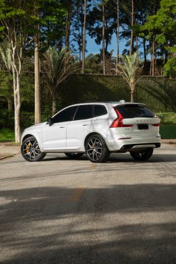Beyaz Volvo XC60 Polestar arka üç çeyreklik, Melez SUV asfalt yolda ağaçlarla park etmiş.