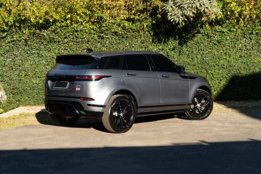 Gri Range Rover Evoque üç çeyreklik, lüks SUV yeşil duvarın ve ağaçların önüne park edilmiş.