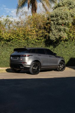 Gri Range Rover Evoque üç çeyreklik, lüks SUV yeşil duvarın ve ağaçların önüne park edilmiş.