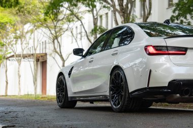 Beyaz BMW M3 yarışması arka manzara, lüks spor sedan yeşil ağaçların altında ıslak asfalta park edilmiş.