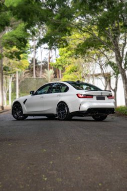Beyaz BMW M3 yarışması. Arka manzara. Üç çeyrek, asfalt yolda yeşil ağaçlarla kaplı spor sedan.
