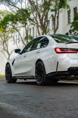 Beyaz BMW M3 yarışması arka manzara, lüks spor sedan yeşil ağaçların altında ıslak asfalta park edilmiş.