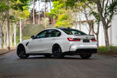 Beyaz BMW M3 yarışması. Arka manzara. Üç çeyrek, asfalt yolda yeşil ağaçlarla kaplı spor sedan.