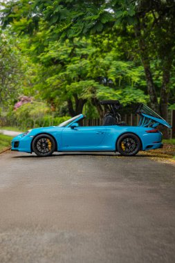 Mavi Porsche 911 Targa 4 GTS yan görüntüsü yarı açık, bahçe düzeninde lüks bir spor araba.
