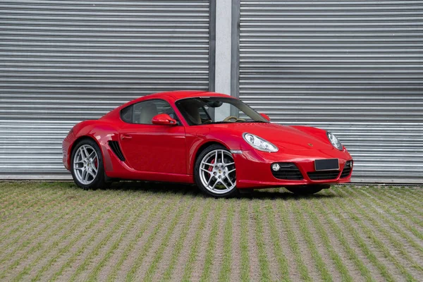Kırmızı Porsche Cayman S önde üç çeyrek manzara, lüks spor araba endüstriyel kapının önünde.