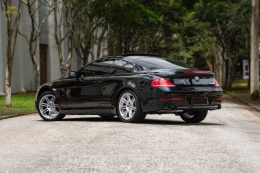 Siyah BMW 650i arka üç çeyrek görüş, boş yolda lüks coupe ağaçlarla çevrili