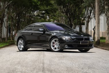 Siyah BMW 650i ön manzara üç çeyrek, lüks sedan ağaçlı yolda bulutlu bir günde park edilmiş.