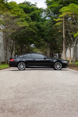 Siyah BMW 650i yan görünüm, yemyeşil ağaçlarla dolu boş yolda lüks coupe.
