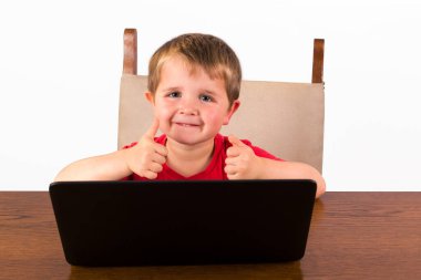 4 yaşında tatlı bir çocuk dizüstü bilgisayarın arkasında oturmuş kameraya bakıp gülümserken iki parmağını kaldırıyor. beyaz arkaplanda izole