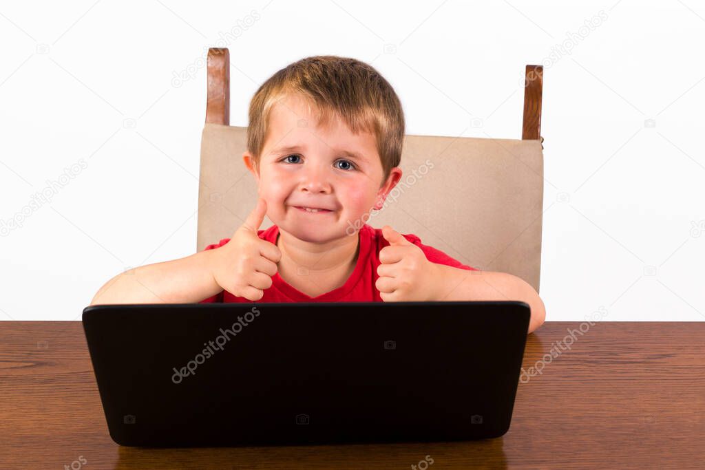 Cute 4 year old toddler sitting behind laptop computer giving two thumbs up while looking at the camera and smiling. isolated on white background