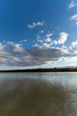 Bulutlar güneşli bir öğleden sonra bir nehrin üzerindeki gökyüzünde toplanır. Su bulutları ve mavi gökyüzünü yansıtır. Nehir kenarındaki ağaçlar yeşile boyanıyor..