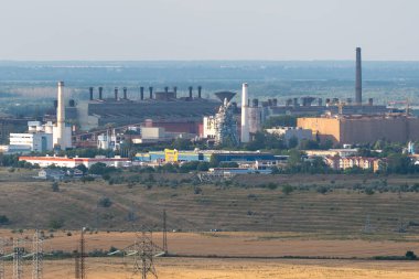 Manzarada fabrikalar ve uzun bacalar mevcuttur. Binalar, açık alanların yakınındaki bir sanayi bölgesinin bir kısmını oluşturuyor. Gökyüzü açık ve sahne yoğun bir çalışma ortamı gösteriyor..