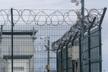 Dikenli telleri olan bir güvenlik çiti, şehir merkezindeki bir tesisi kapatıyor. Çitin arkasındaki yapılar daha büyük bir kompleksin parçası gibi görünüyor. Gözetleme ekipmanları görünür..